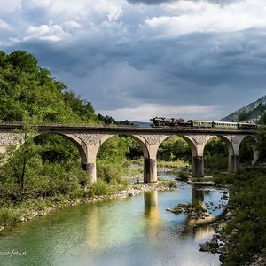 Fotografija železniškega mostu, po katerem vozi muzejska parna lokomotiva. Pod mostom teče reka Soča.